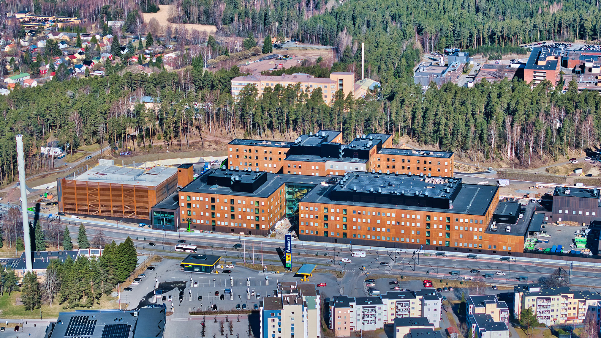 Aerial view of a large modern building complex surrounded by trees, car parks, and roads, with a mix of residential and commercial areas in the background.
