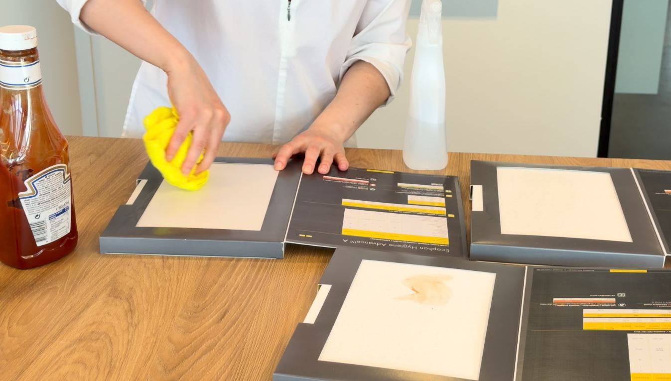 A person cleans a stain from a laminated surface using a yellow cloth. There are other stained surfaces, a spray bottle, and a ketchup bottle on the wooden table.
