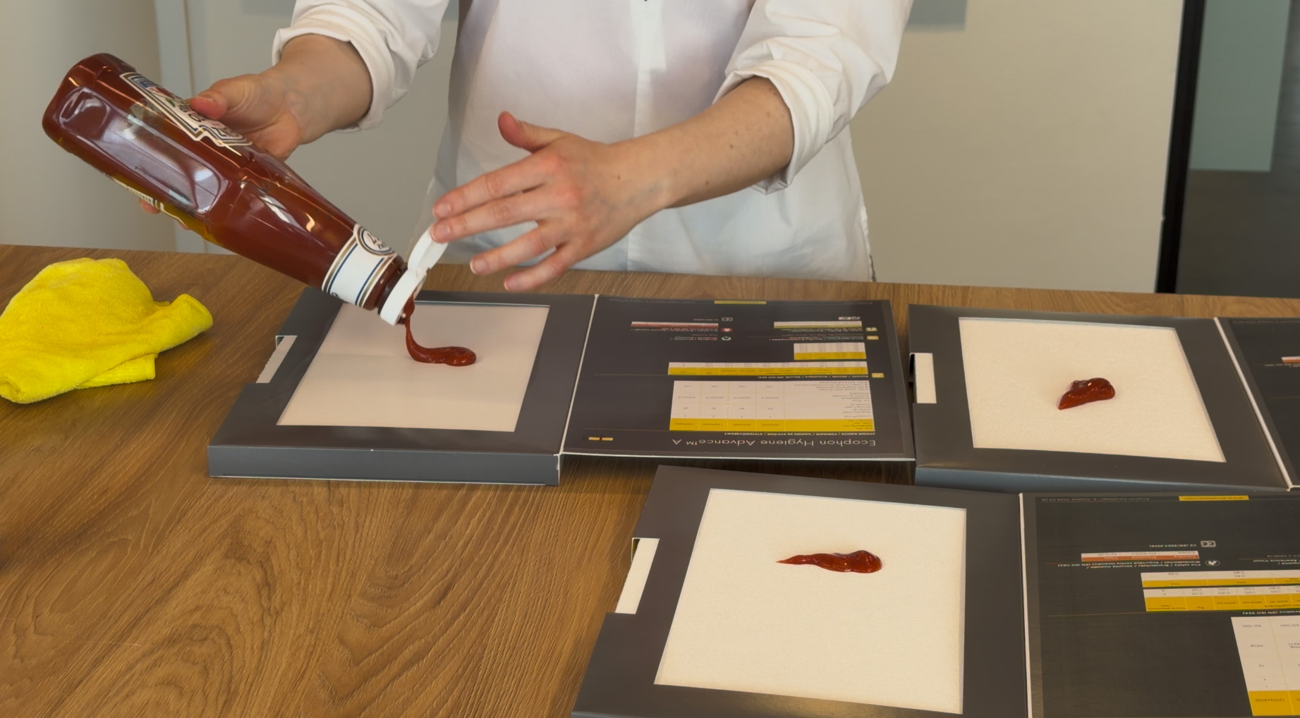 A person in a white shirt pours tomato sauce from a bottle onto a white square surface, with other similar squares and a yellow cloth on a wooden table.