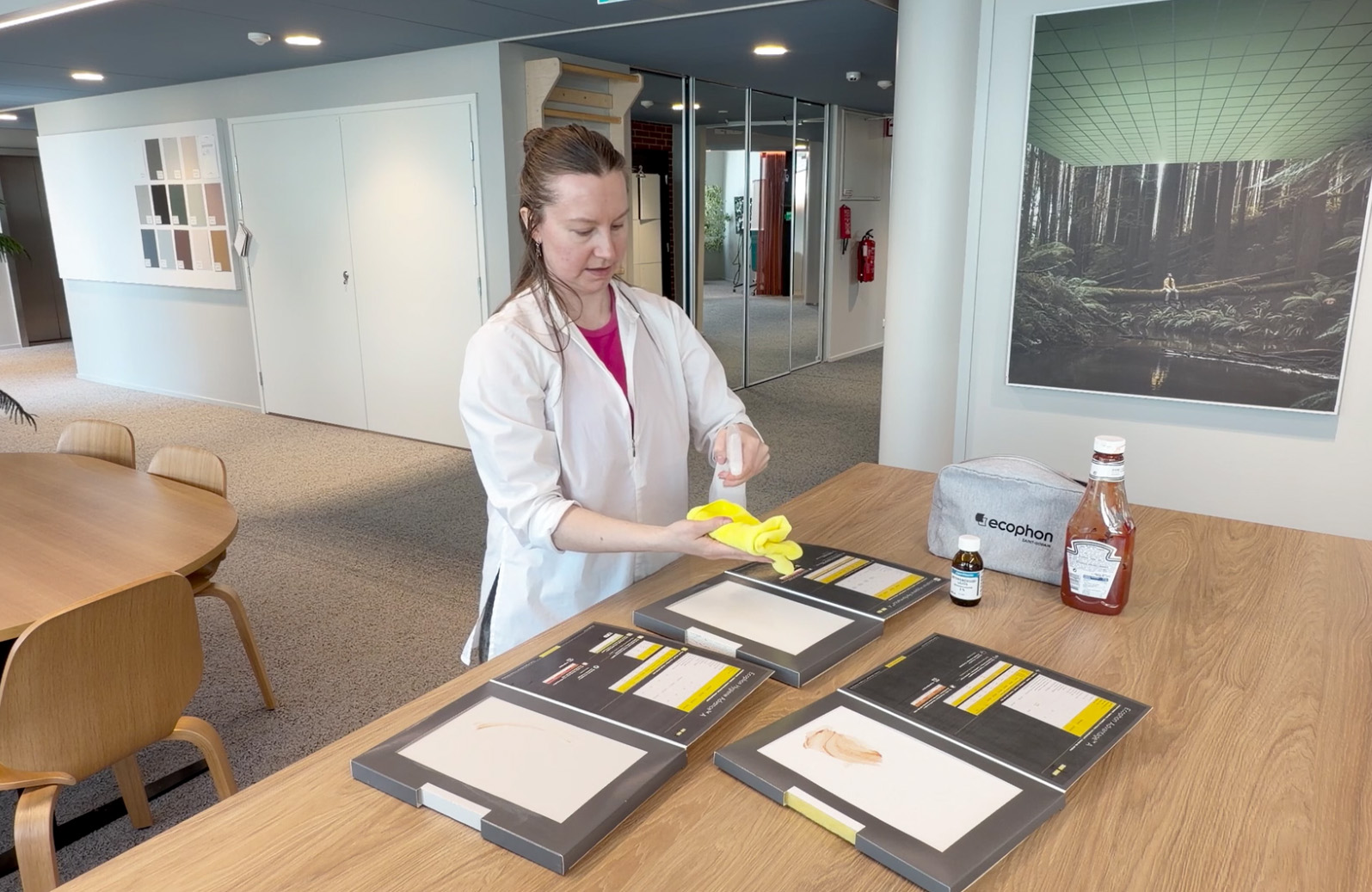 A woman in a lab coat wipes a yellow cloth on a test panel on a table, with a bottle of tomato sauce, a grey bag, and swab kits nearby in a modern office space.