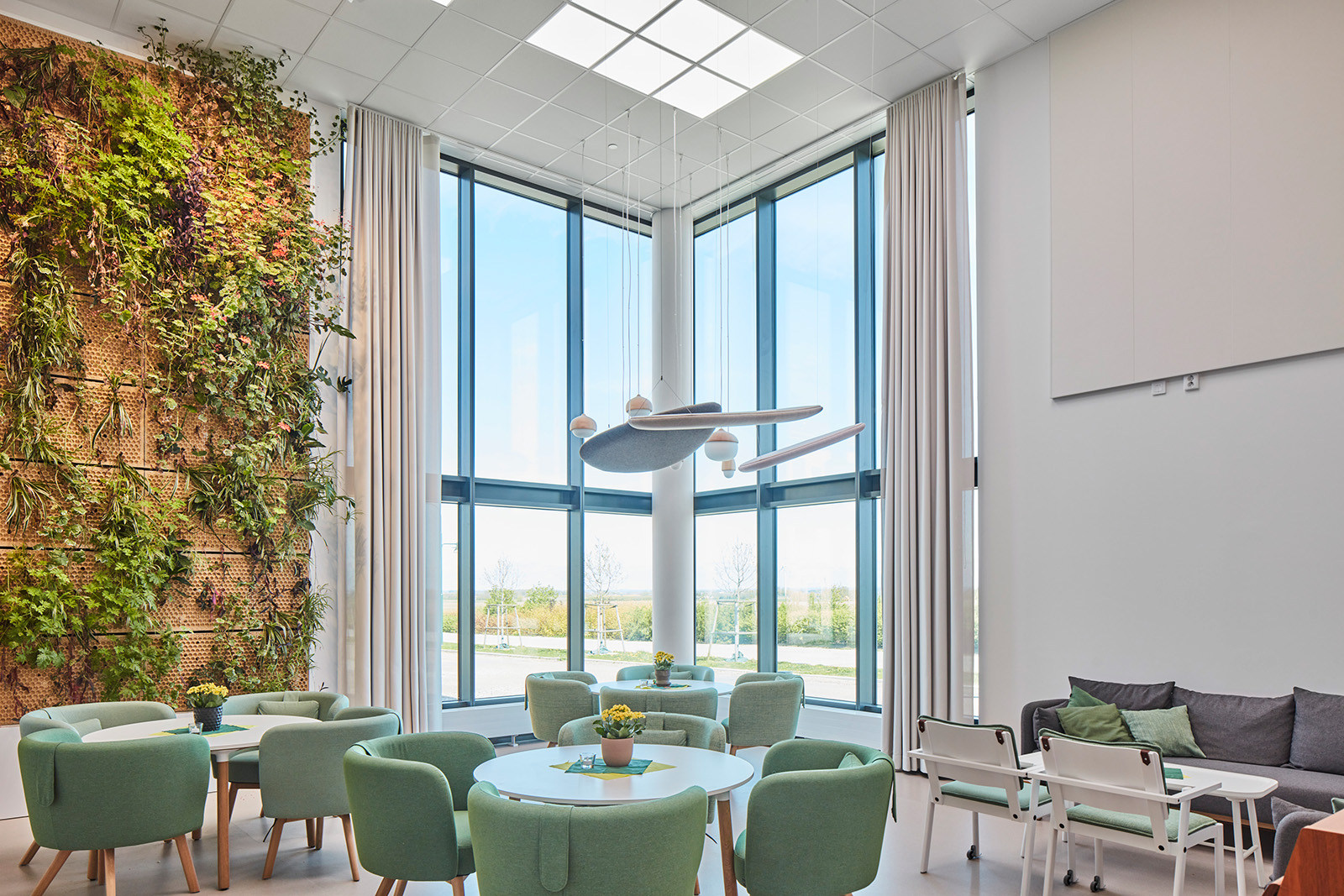 A modern and bright room with large windows providing ample natural light. The room features a living green wall with various plants on the left side, adding a touch of nature indoors. In the center, there are round tables surrounded by mint green chairs with a yellow floral centerpiece. Above the table hangs an artistic light fixture with multiple round lights at different heights. To the right, there is a cozy seating area with grey sofas and chairs around a low coffee table. The overall atmosphere is clean, refreshing, and designed for relaxation or casual meetings.