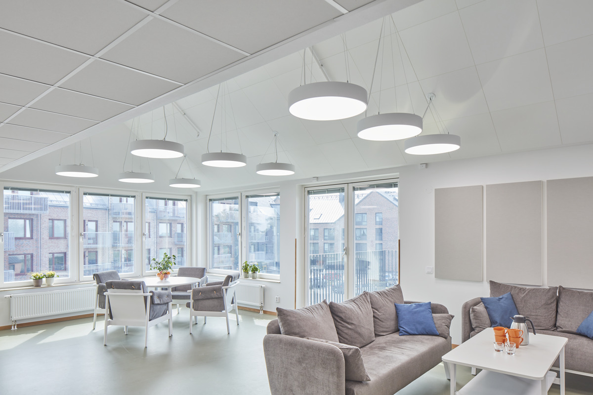 A modern, brightly lit break room with large windows offering an outside view. The room features a plush grey sofa with blue cushions, several white tables paired with chairs, and circular overhead lighting fixtures. The walls are adorned with white acoustic panels, creating a clean, minimalist aesthetic conducive to relaxation.
