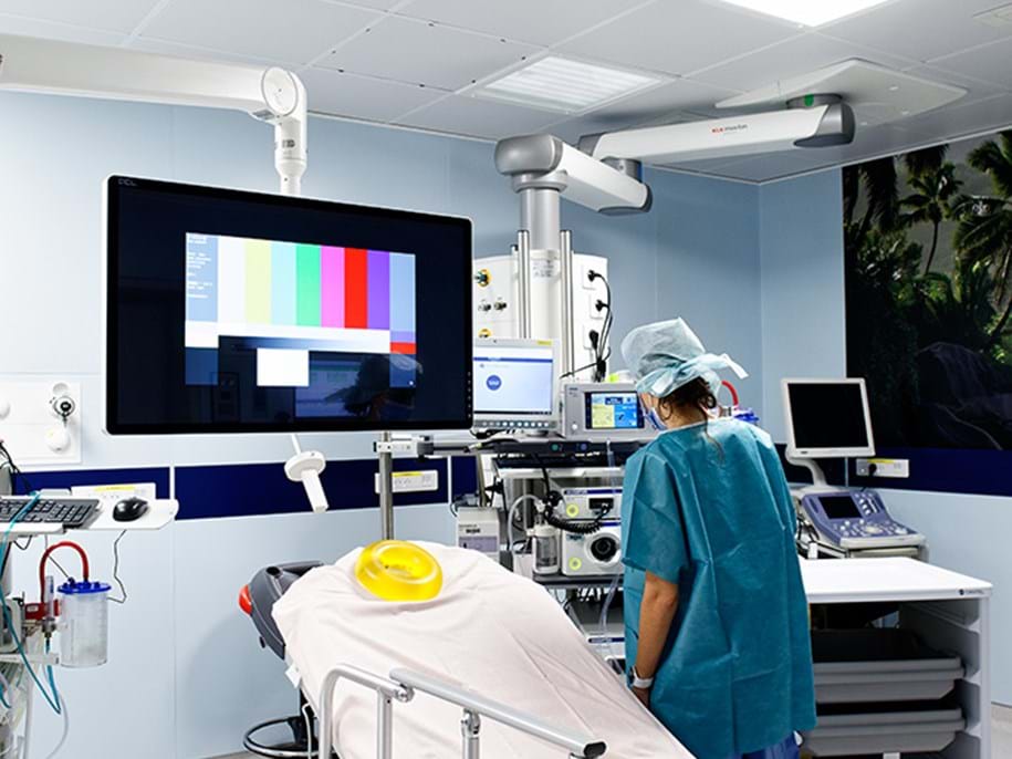 A medical professional in scrubs and a hair cover stands beside equipment in a modern hospital operating room with a covered bed and a large screen displaying a colour test pattern.