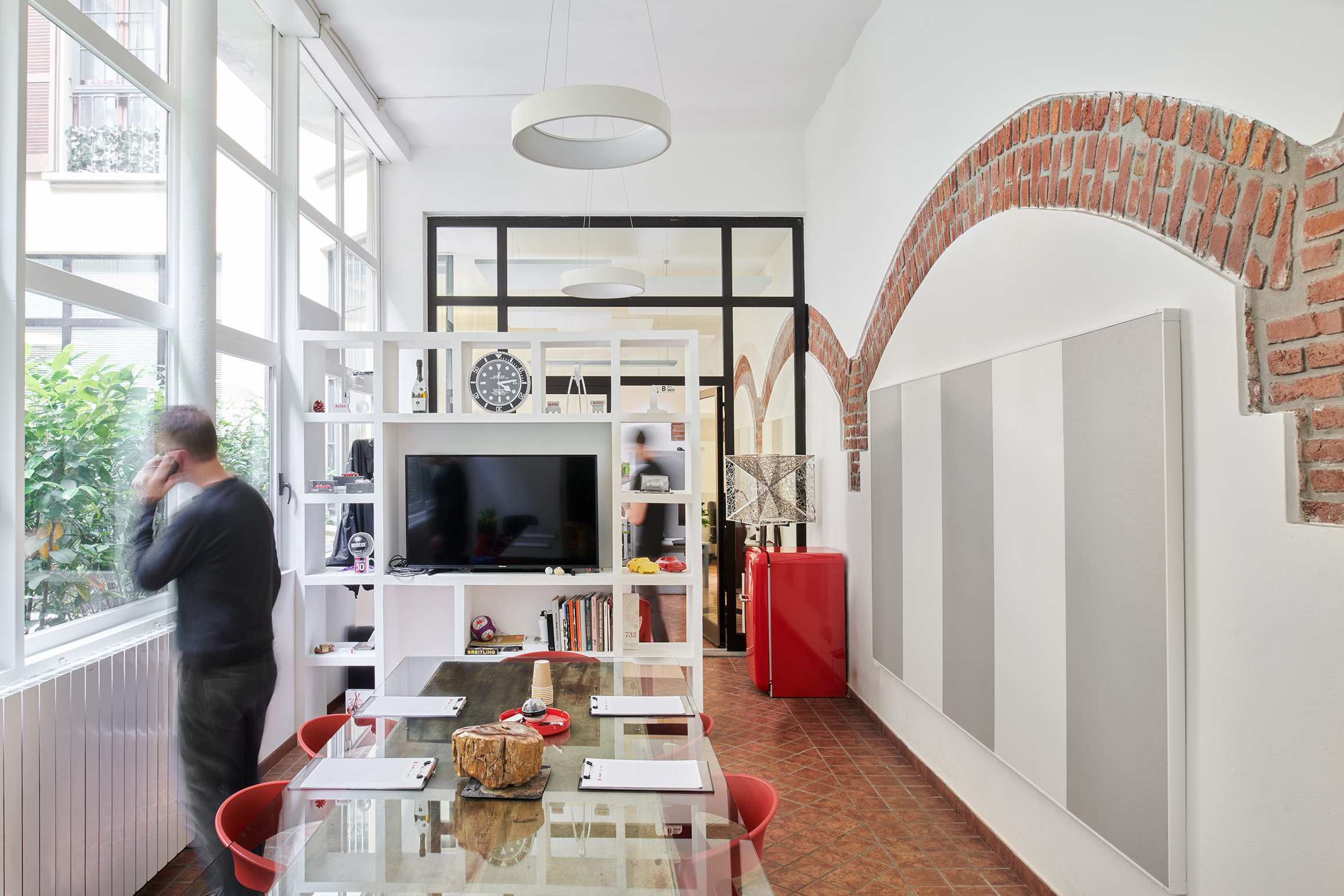 A modern open-plan office with a large glass table and red chairs. A black TV is mounted on a white shelf with various decor items. A person stands by tall windows with a view outside. Exposed brick arches adorn white walls, and a red fridge is in the corner.