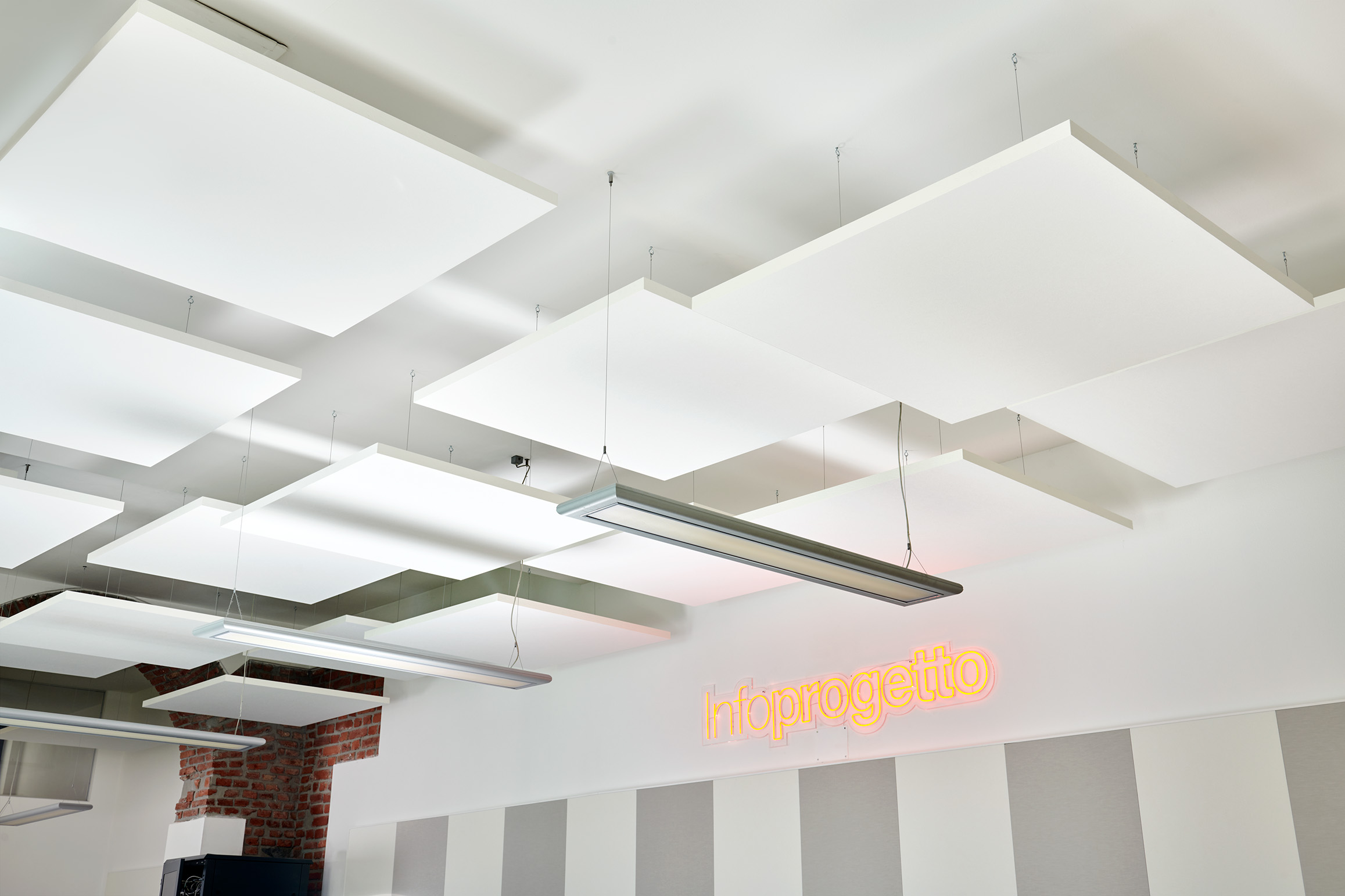 Ceiling with rectangular acoustic panels and hanging lights in a modern room. A sign on the wall reads Infoprogetto in orange neon. Red brick accents and white wall panels are visible.