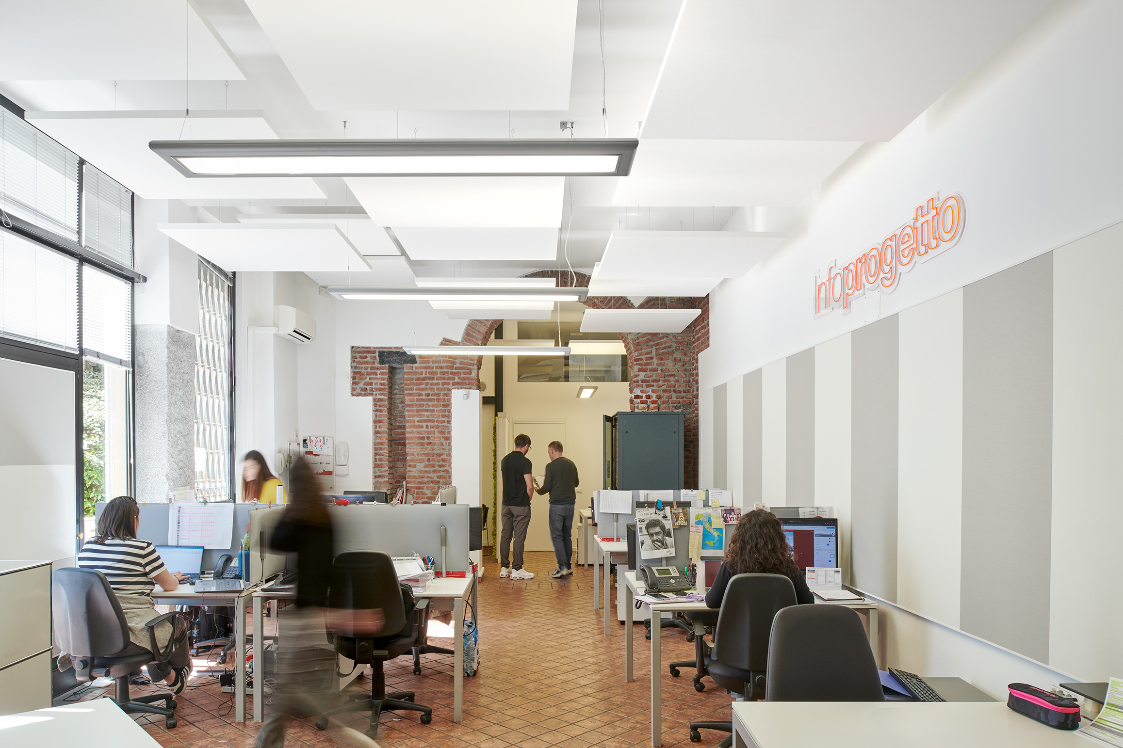 An open office space with people working at desks equipped with computers. The room has white walls, brick accents, and modern lighting. Two people are standing and talking in the background near a blue door.