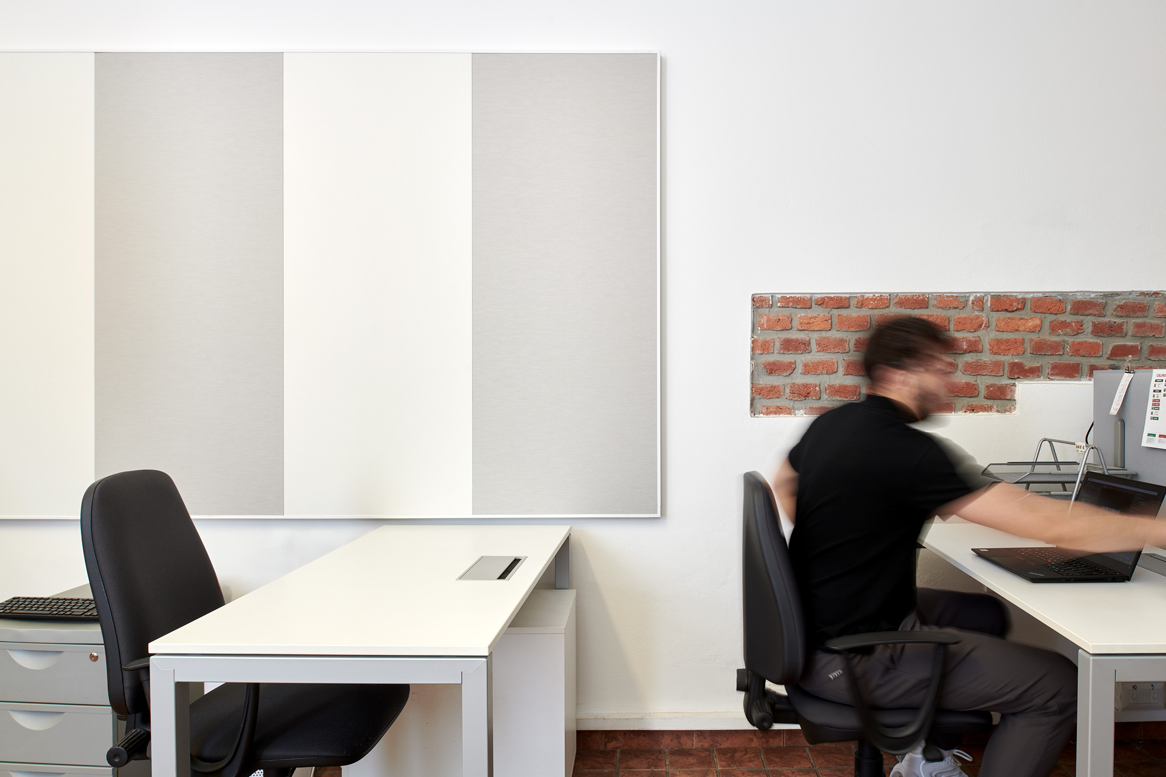 A person in a black shirt moves quickly in a modern office space with white desks and chairs. Behind, a brick and white wall is visible, along with vertical canvas panels. The scene appears dynamic with a sense of motion.