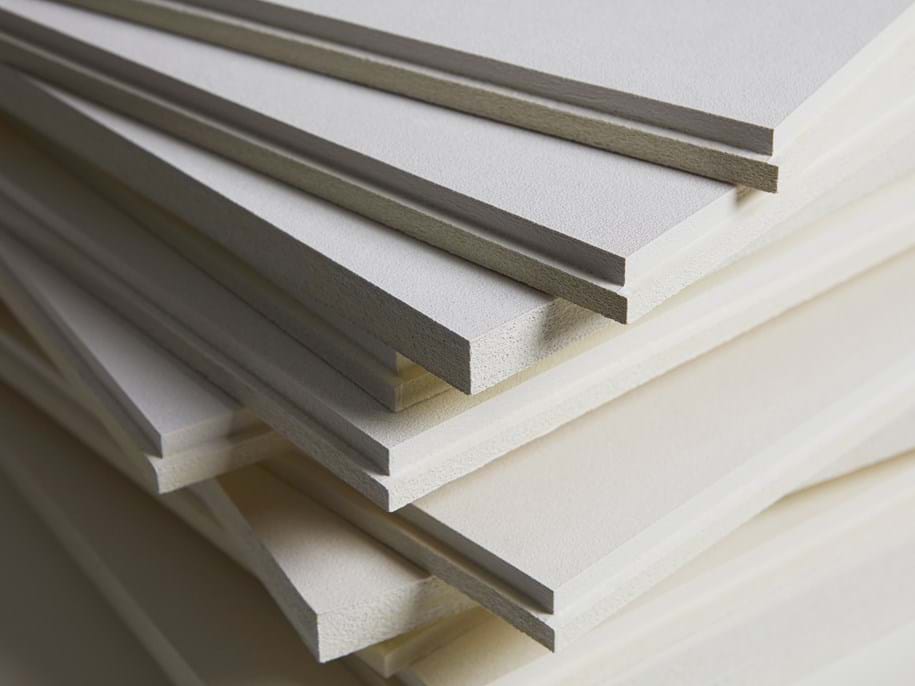 A close-up of multiple white acoustic boards stacked on top of each other, showing their thin edges and layered arrangement. The smooth surfaces and precise cuts are visible, creating a geometric pattern.