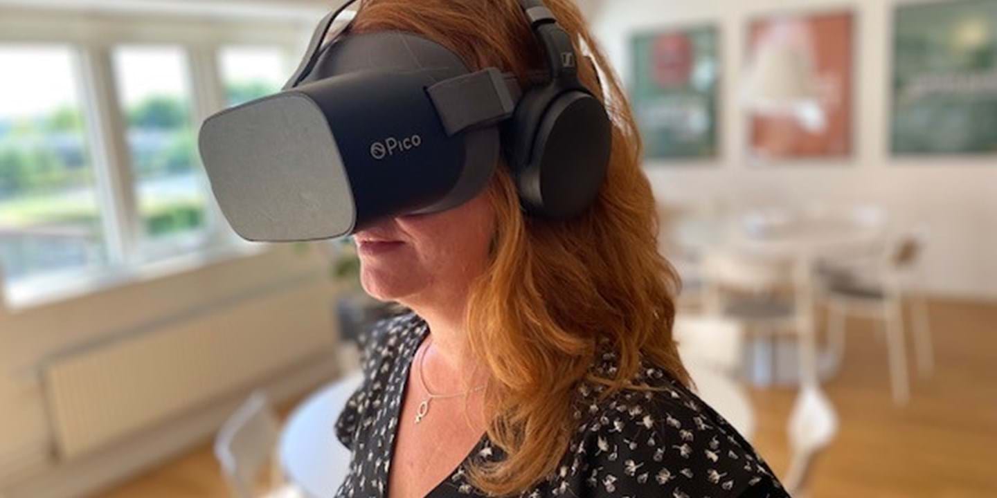 Eine Frau mit langen kastanienbraunen Haaren trägt ein Pico Virtual-Reality-Headset und Kopfhörer. Sie befindet sich in einem hellen, modernen Raum mit Holzboden und weißen Möbeln. An den Wänden im Hintergrund sind unscharfe Kunstwerke zu sehen.