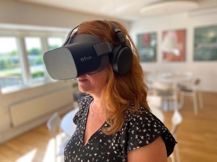 A woman with long auburn hair is wearing a Pico virtual reality headset and headphones. She is in a bright, modern room with wooden floors and white furniture. Blurred artwork is displayed on the walls in the background.