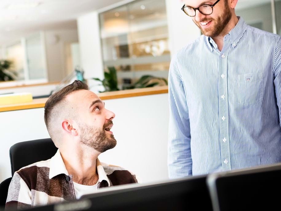 Two men with beards are in an office setting. One is sitting at a desk, smiling up at the other, who is standing and wearing glasses. They appear to be having a friendly conversation. Several computer screens and office decor are visible.