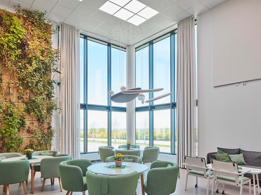 A modern, spacious room with large floor-to-ceiling windows allowing ample natural light. The room features a vertical garden on one wall. There are round tables surrounded by light green upholstered chairs, and a cozy seating area with a grey sofa and white chairs. The room has a contemporary light fixture hanging from the acoustic ceiling with biocentric lighting.
