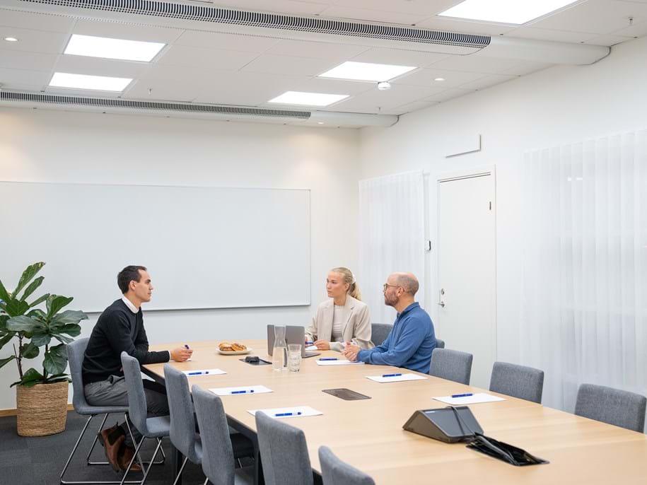 A modern meeting room with a long wooden conference table surrounded by grey upholstered chairs. Three people are seated at the table engaged in a discussion, with notepads and pens in front of them. The room features a large whiteboard on the wall, a potted plant for decoration, and bright overhead lighting. The ceiling features acoustic panels and biocentric lighting.