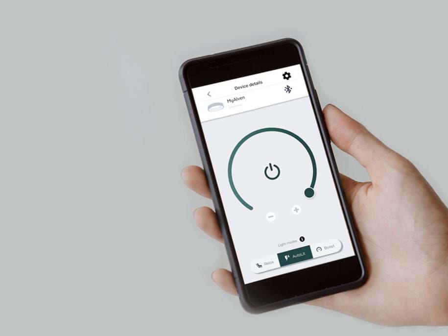 A hand holding a smartphone displaying a control app interface. The screen shows a circular power button with options to adjust settings, and below it are buttons labeled "Relax," "AutoLit," and "Boost." The background is plain and light grey, emphasising the focus on the smartphone and the app interface.