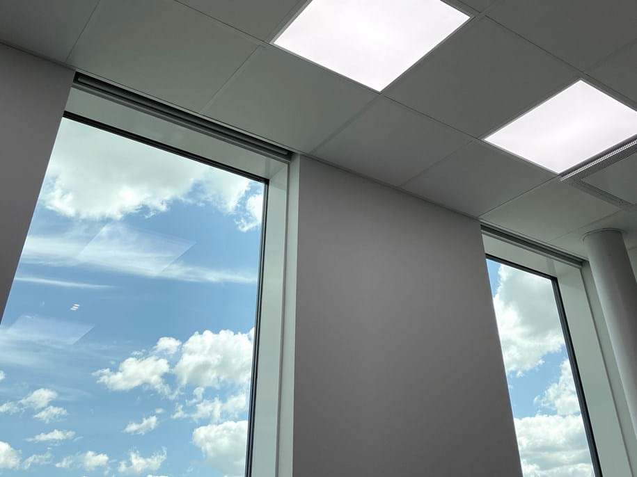 A view of a room with large windows showing a clear blue sky with scattered clouds, and biocentric ceiling lights turned on.