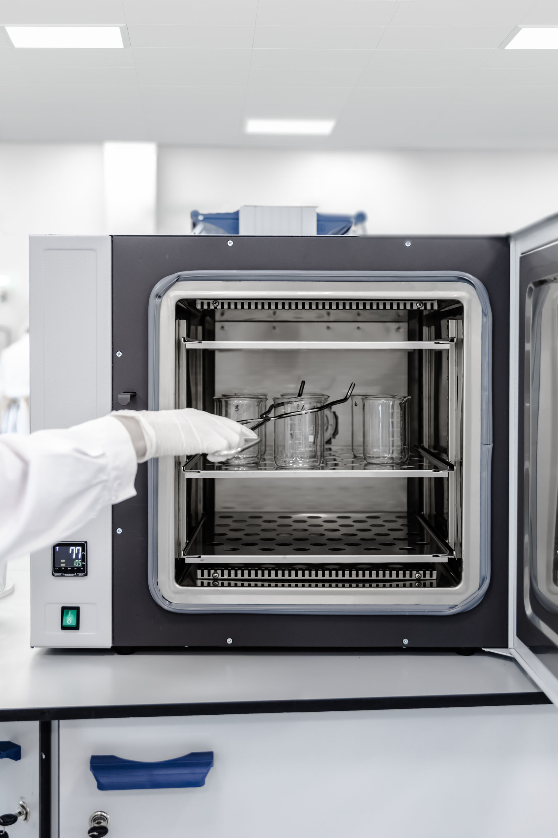 A gloved hand places a glass beaker inside a laboratory oven with several other beakers on shelves. The oven door is open, displaying a digital temperature readout and a clean, modern lab environment.