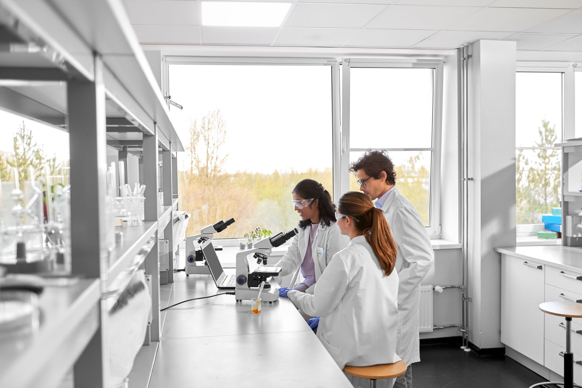 Three scientists wearing lab coats and goggles work together at a laboratory bench, using a microscope and a laptop, with large windows in the background letting in natural light.