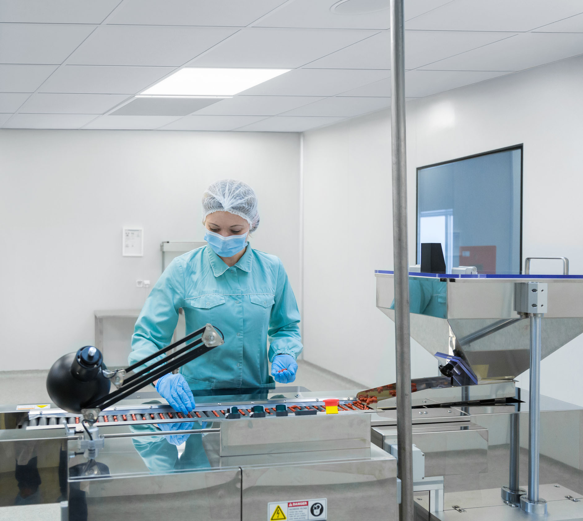 A worker in protective clothing, a hairnet, and a mask inspects pharmaceutical products on a conveyor belt in a sterile, modern laboratory environment.