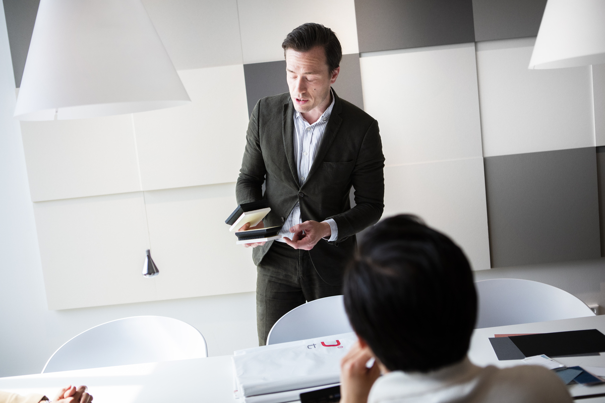 A man in a suit stands at a table, holding notebooks and speaking to colleagues seated in a modern office with geometric wall panels and white hanging lamps.