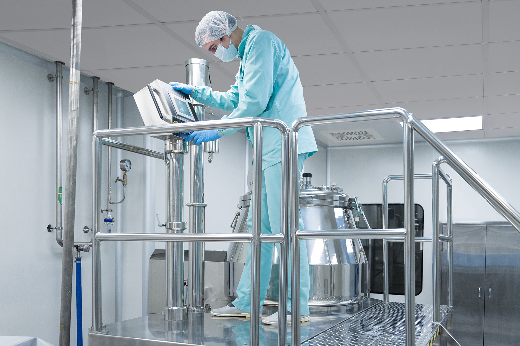 A person in protective clothing, gloves, a mask, and a hairnet operates equipment in a clean, modern laboratory or pharmaceutical facility, standing on a metal platform next to a large stainless steel tank.