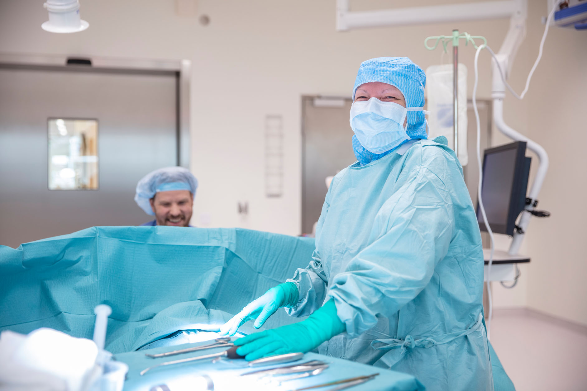 A surgeon wearing a surgical mask, gown, gloves, and cap stands at an operating table with surgical instruments, while another medical professional in theatre scrubs is in the background, both in a brightly lit operating theatre.