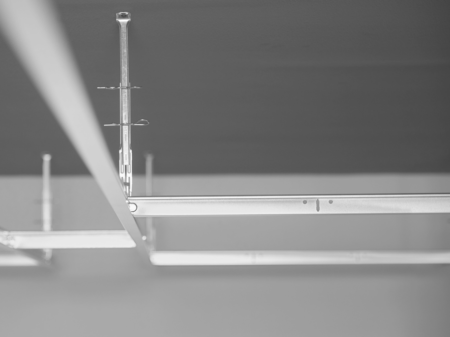A black-and-white photo showing a close-up view of metal ceiling grid framework, with suspension wires attached to a dark ceiling above and a blurred background below.