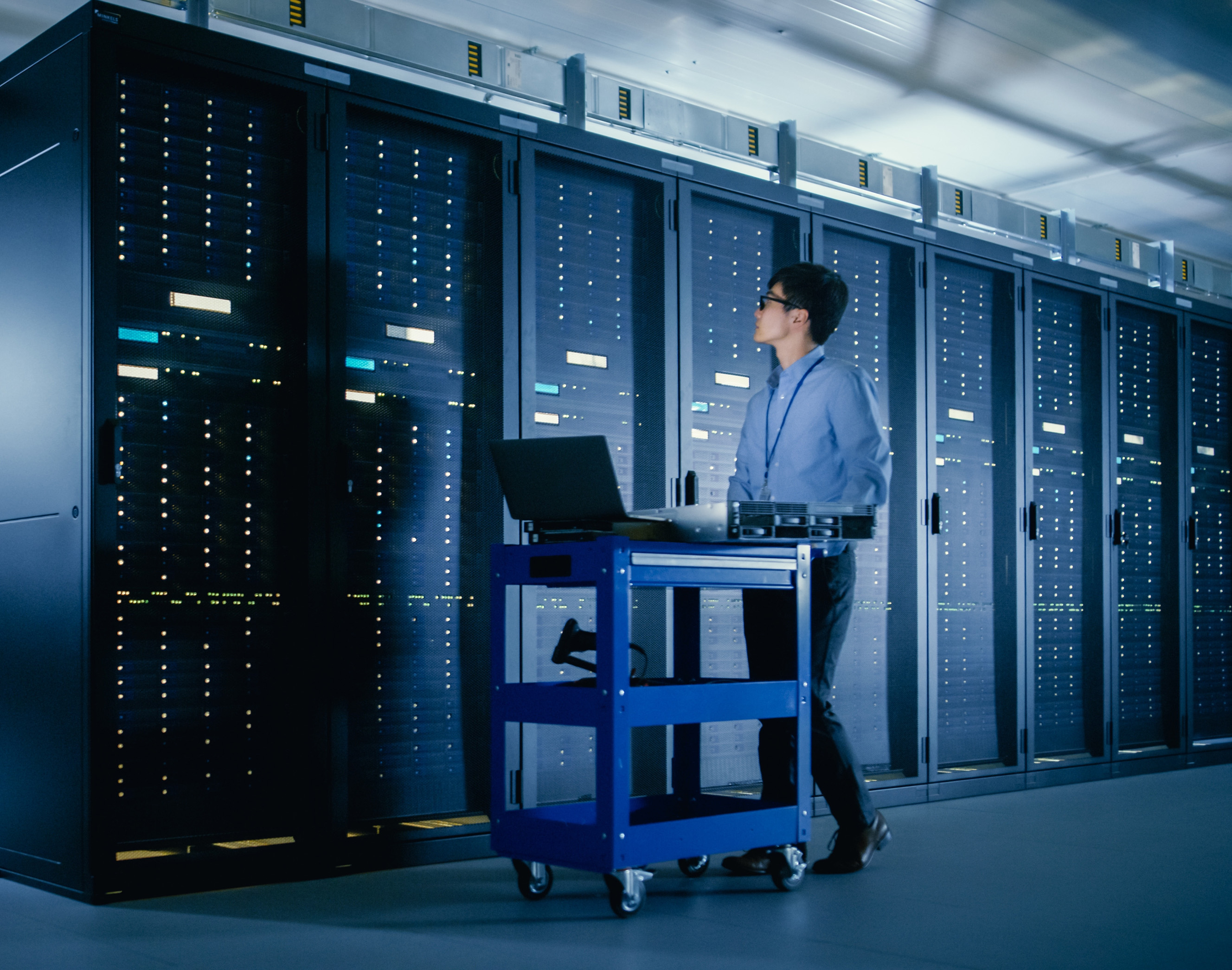 A person stands in a server room, looking at rows of server racks whilst working on a laptop placed on a blue rolling trolley. The environment is brightly lit and filled with computer equipment.