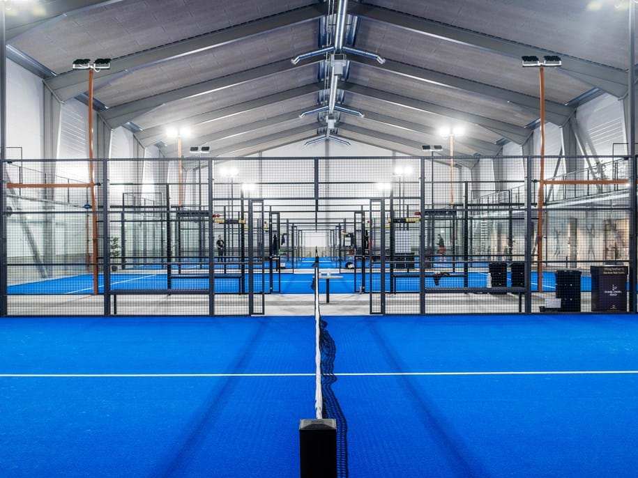 Ein großer Indoor-Padel-Court mit hellblauem Boden und einer hohen, gewölbten Decke. Das Bild zeigt mehrere nebeneinander aufgereihte Courts, umgeben von Glaswänden und Metallnetzen, beleuchtet von Oberlichtern.