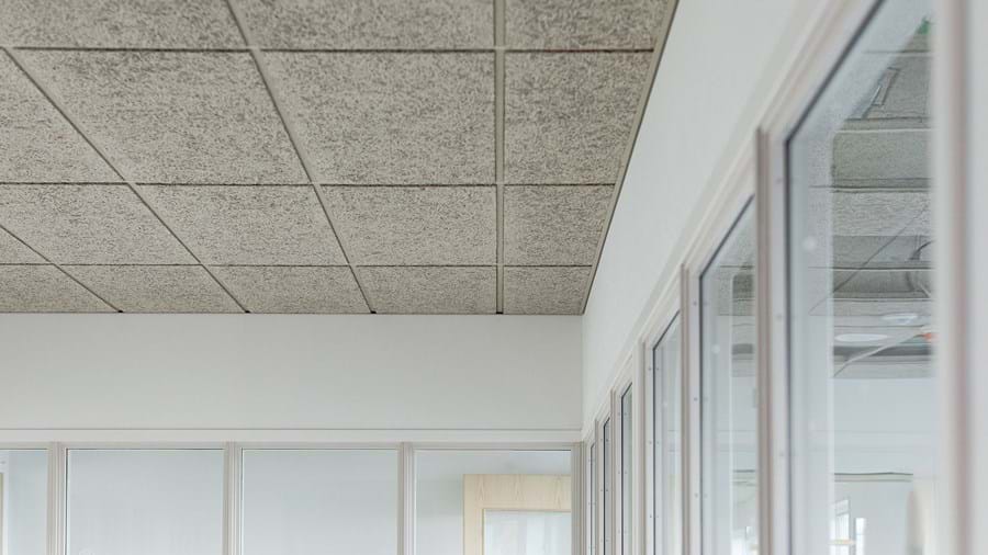 Eine minimalistische Büroeinrichtung mit Schwerpunkt auf der Decke, die aus grauen Akustikfliesen aus Holzwolle besteht. Die Wände sind weiß, und es gibt große Fenster und eine Holztür an der Seite. Der Raum wirkt ruhig und gut beleuchtet.