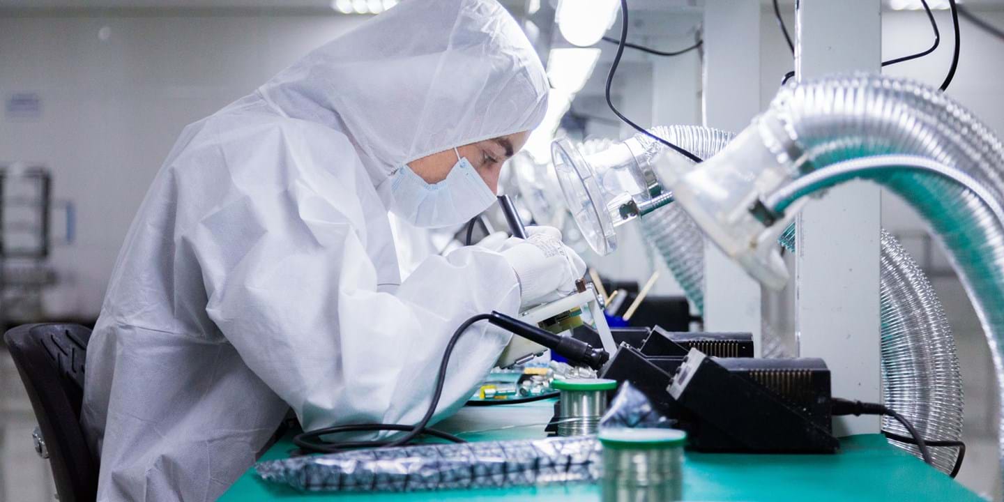 En person i heldragt og maske arbejder med elektroniske komponenter ved en laboratoriebænk, ved hjælp af præcisionsværktøj og omgivet af specialudstyr i et veloplyst, rent miljø.