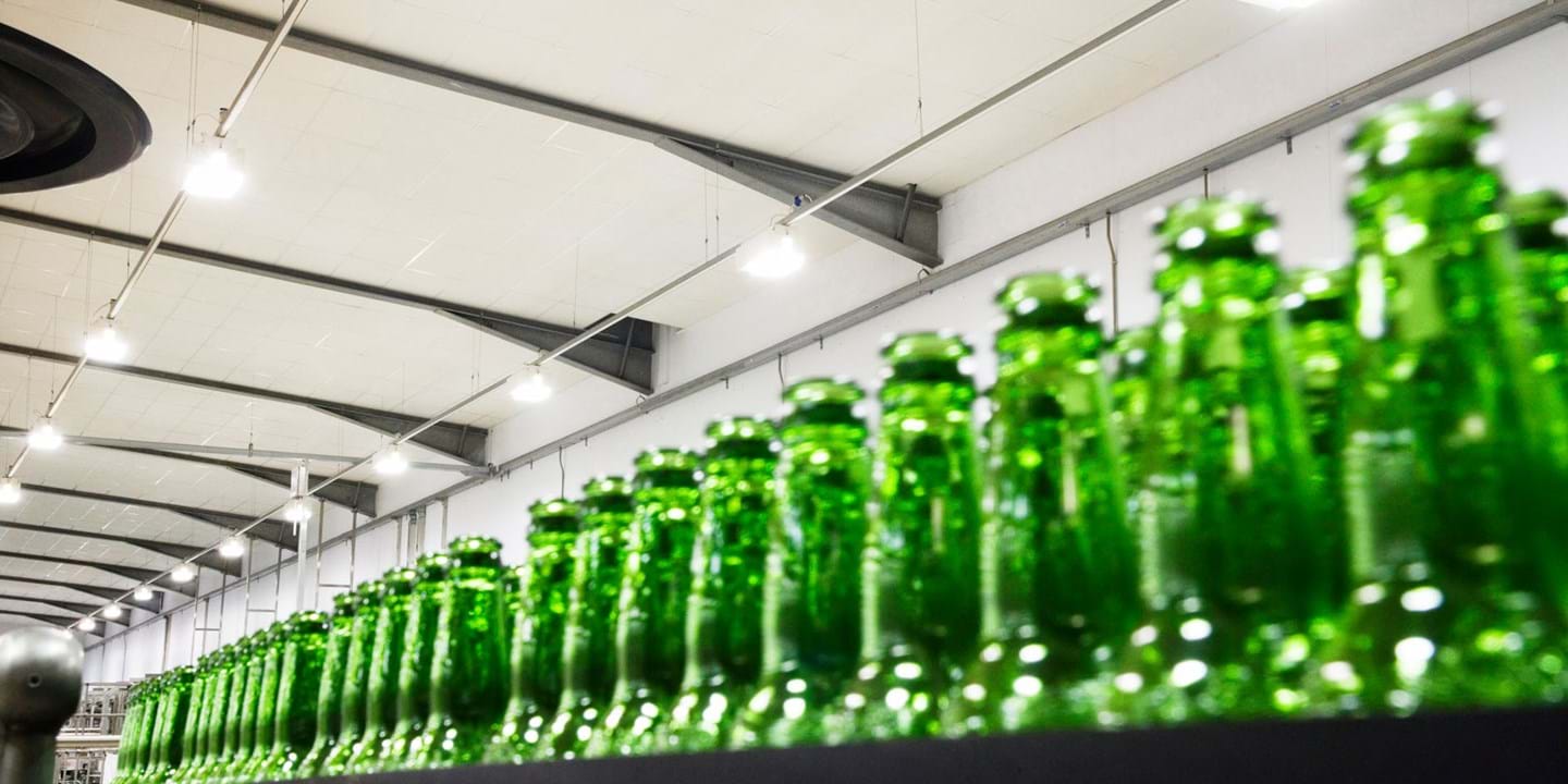 En række tomme grønne glasflasker på et transportbånd i et lyst, industrielt fabriksområde.