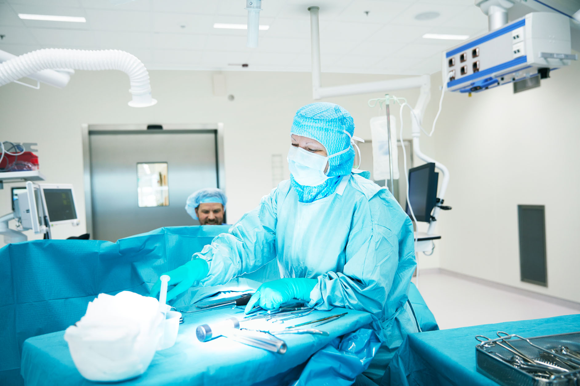 A surgeon in blue scrubs, mask, and gloves performs a procedure in a bright, sterile operating theatre, with another medical professional in the background and surgical instruments arranged on a table.