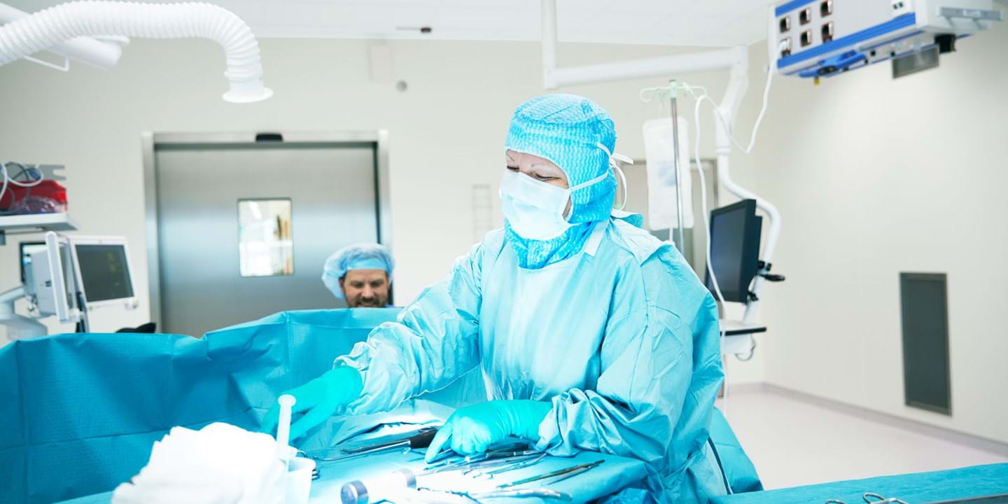 A surgeon in blue scrubs, mask, and gloves performs a procedure in a bright, sterile operating theatre, with another medical professional in the background and surgical instruments arranged on a table.