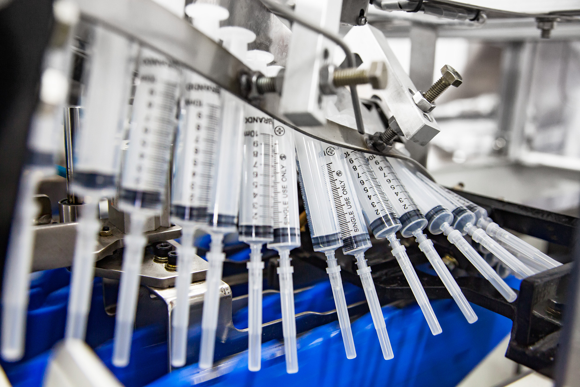 Several plastic syringes are aligned in a row on a production line, held in place by a metal mechanism, in what appears to be a manufacturing or packaging facility.