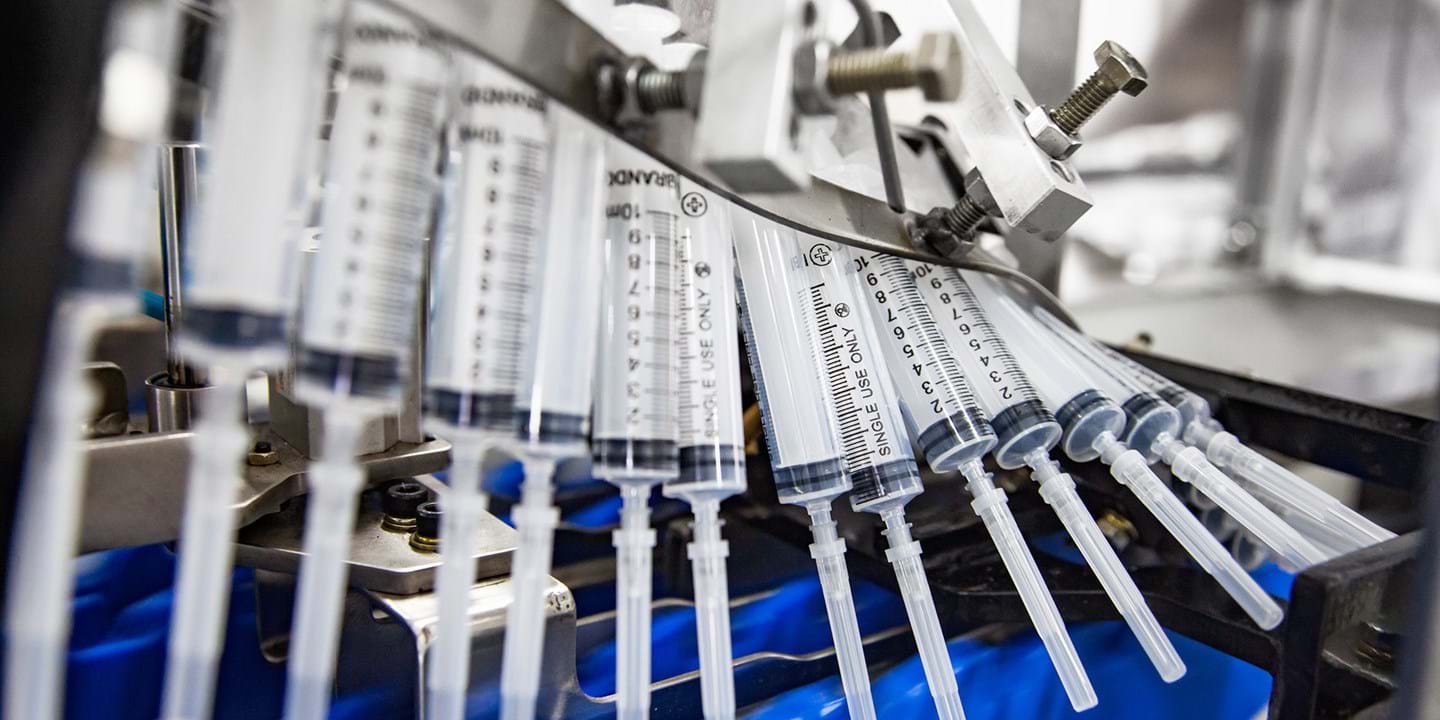 Several plastic syringes are aligned in a row on a production line, held in place by a metal mechanism, in what appears to be a manufacturing or packaging facility.
