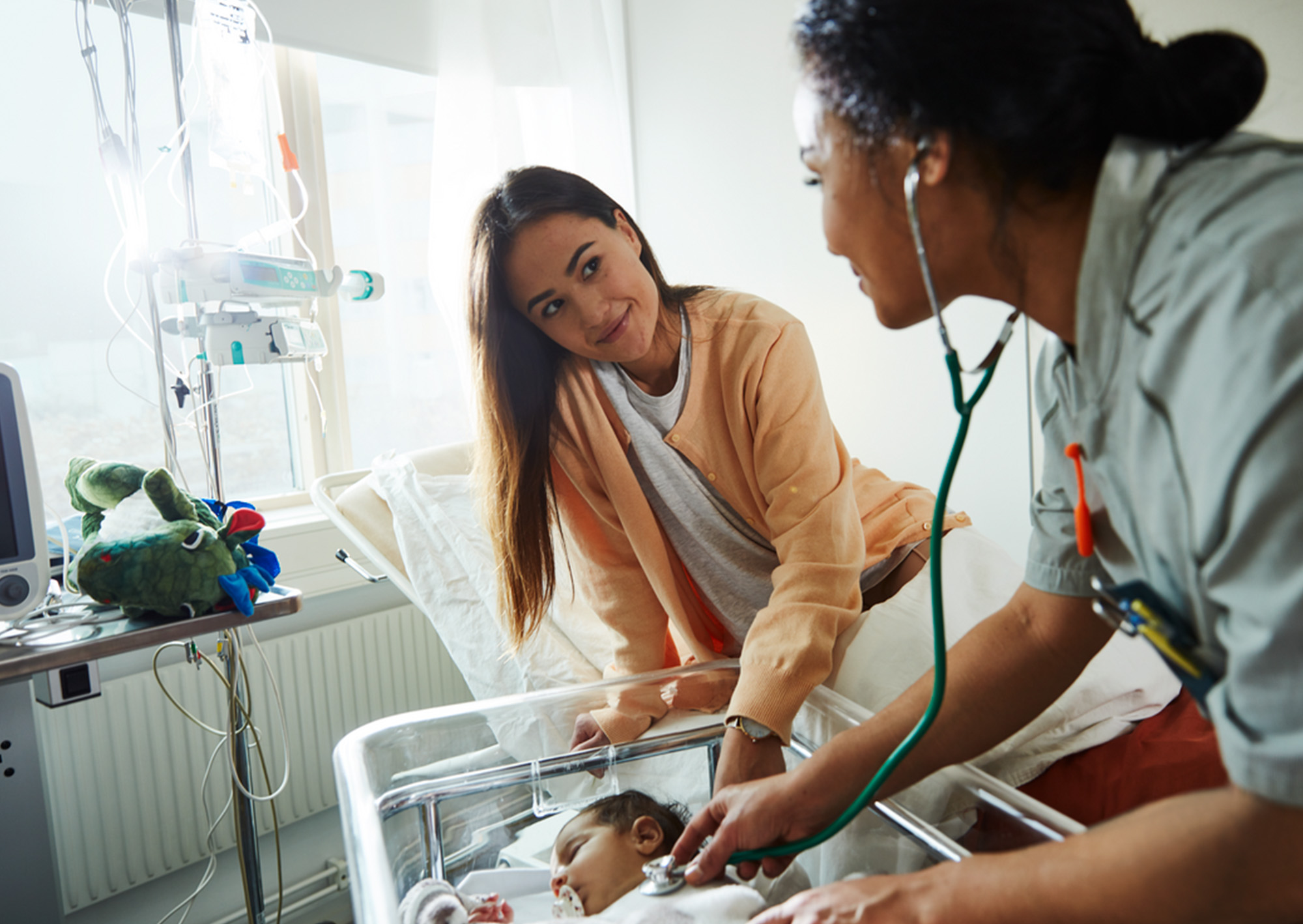 A healthcare worker uses a stethoscope to check a newborn who is lying in a hospital cot, while an adult woman, possibly the mother, smiles and looks on at the baby's bedside.
