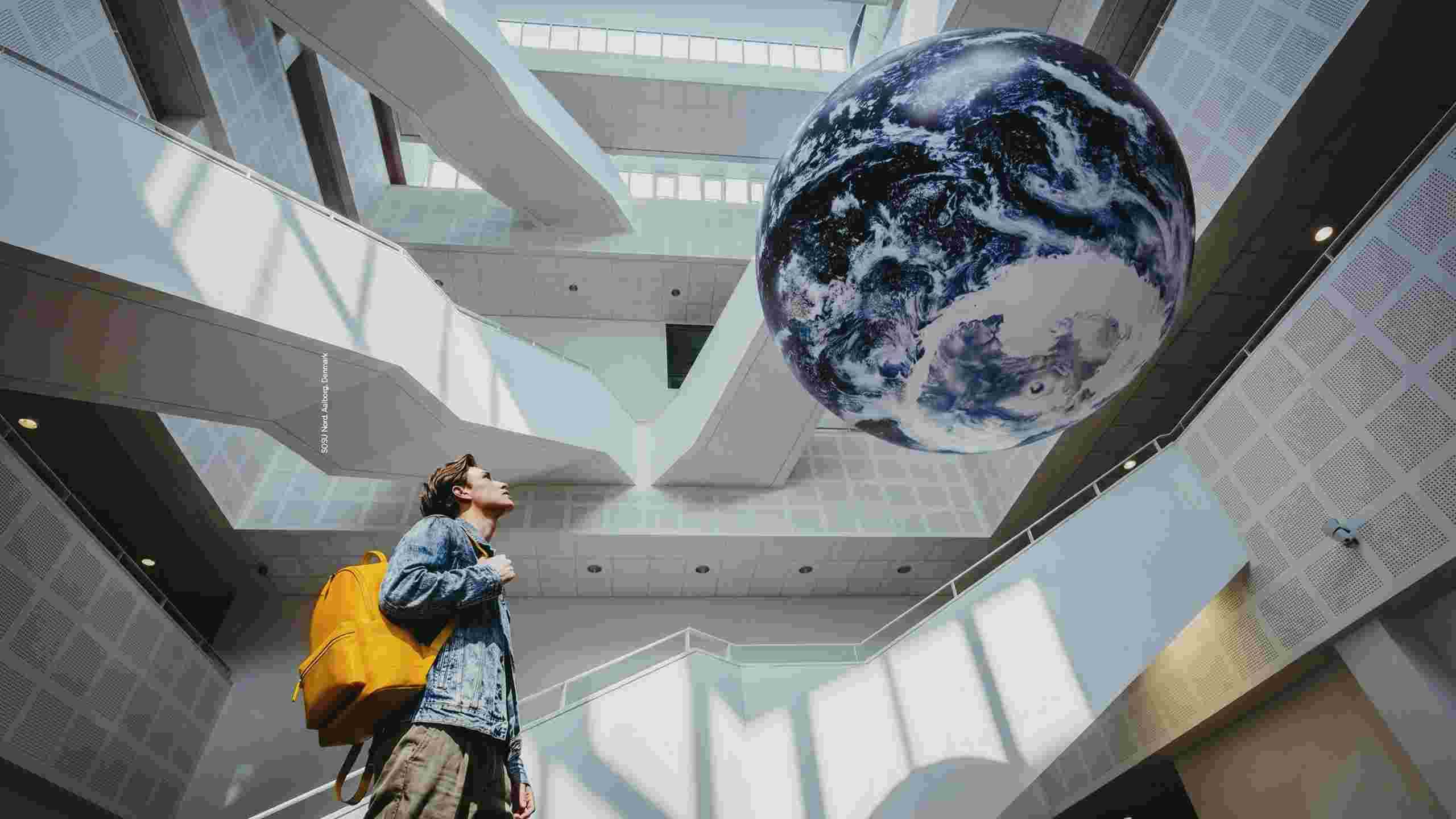 A person with a yellow rucksack looks up at a large globe of Earth suspended in a modern, white architectural space with geometric lines.