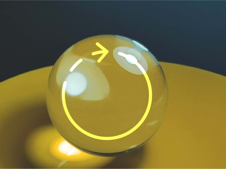 A reflective, transparent sphere with a yellow arrow and circle drawn on it sits atop a yellow surface against a dark background. The arrow points upwards, creating a sense of motion.